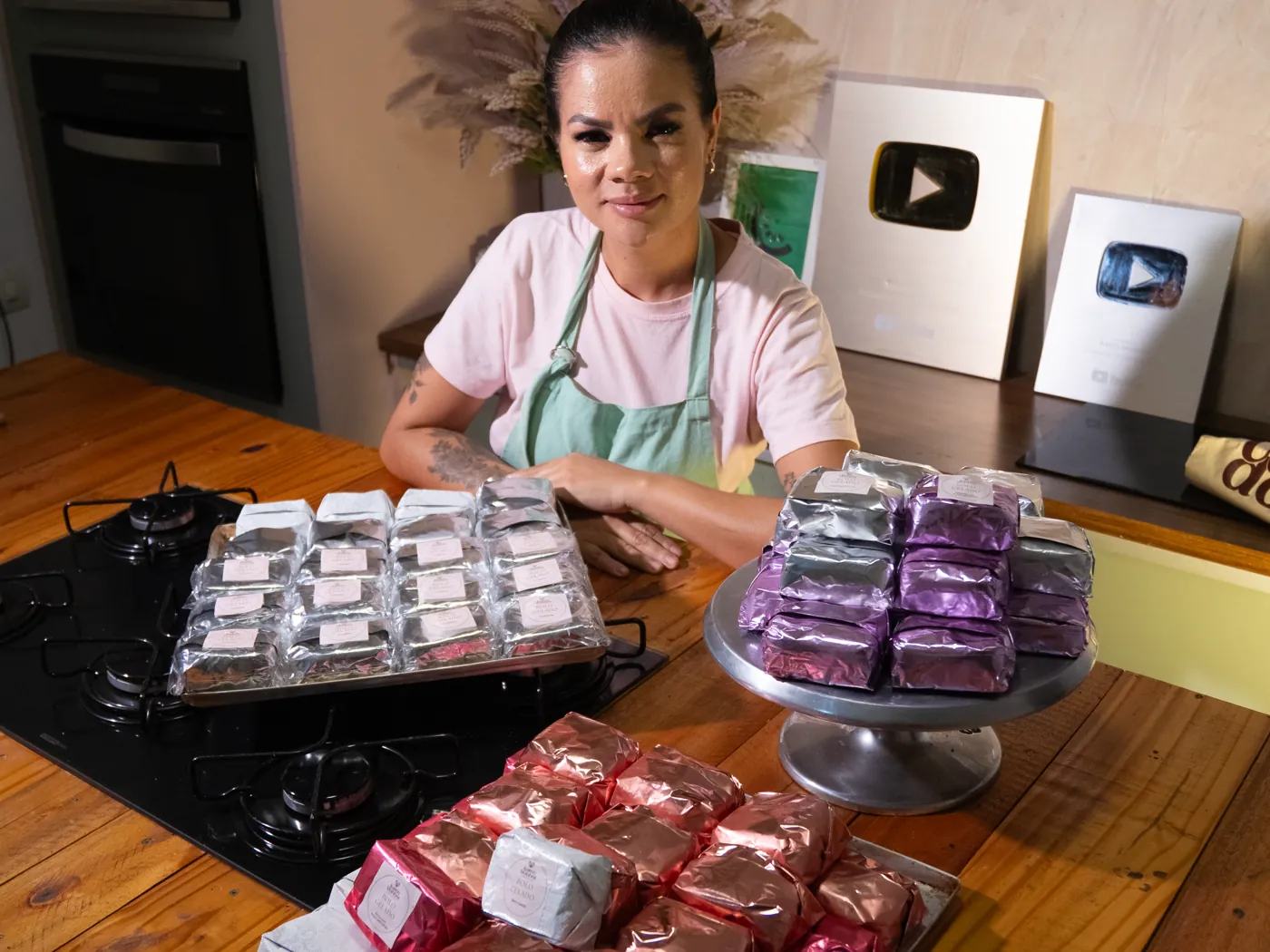 Karen Munhoz atrás de três bandejas de bolos gelados embalados, com as placas do YouTube ao fundo